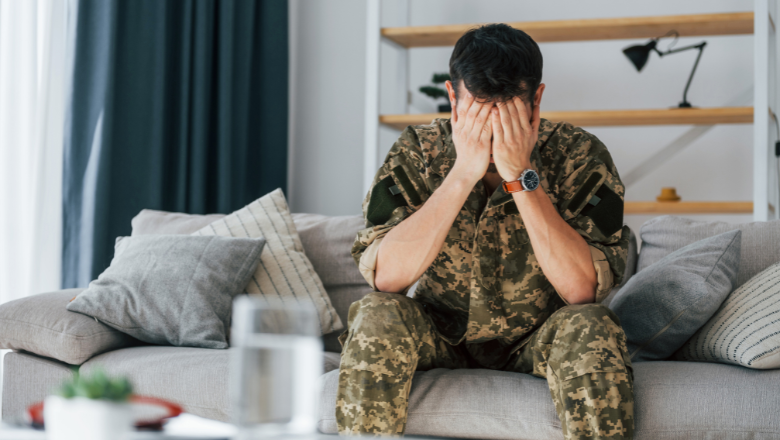 Photo of military personnel in camouflage combat clothing, holding their face in their hands, possibly crying or sad. They are seated on a sofa, probably in a living room.