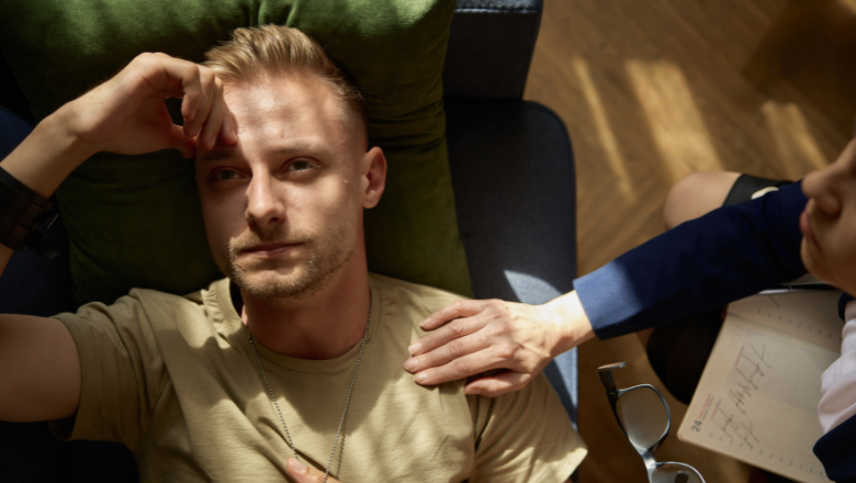 Portrait of a soldier in a therapy setting. His forehead is resting against his hand and he stares into the distance. He is seated in an armchair, and the hand of a therapist is resting on his shoulder for comfort.