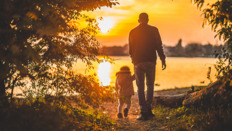 A parent and child holding hands are seen from behind them. They are walking forwards towards the sunset.