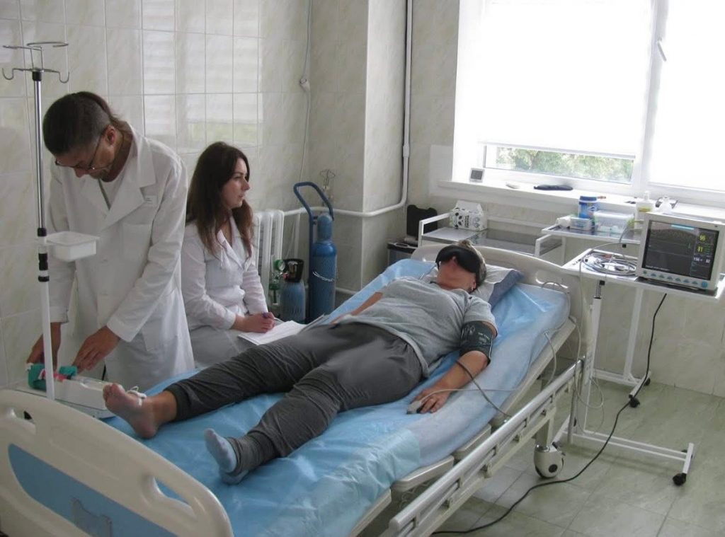 Photo of a patient being treated at Expio Clinic in Kyiv. The patient is lying on a hospital bed with a heart rate monitor and an eyemask on their face to help them relax and gain the most benefit from their session.
To their left, medical staff checking their vitals as well as a therapist can be seen. The overal atmosphere is calm and soft white light is coming through the white blinds on the window.
