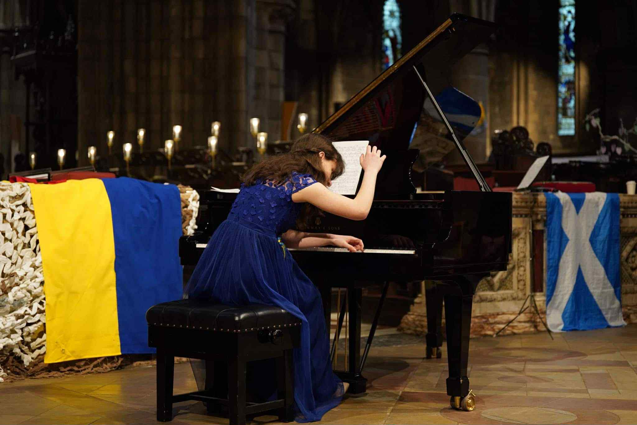 Photo of one of the Intertwined muscians playing the piano during a concert at St Cuthbert's Church in Edinburgh. Her arm is raised as she is about to touch the keyboard. In the background, a Ukrainian and a Scottish flag can be seen.