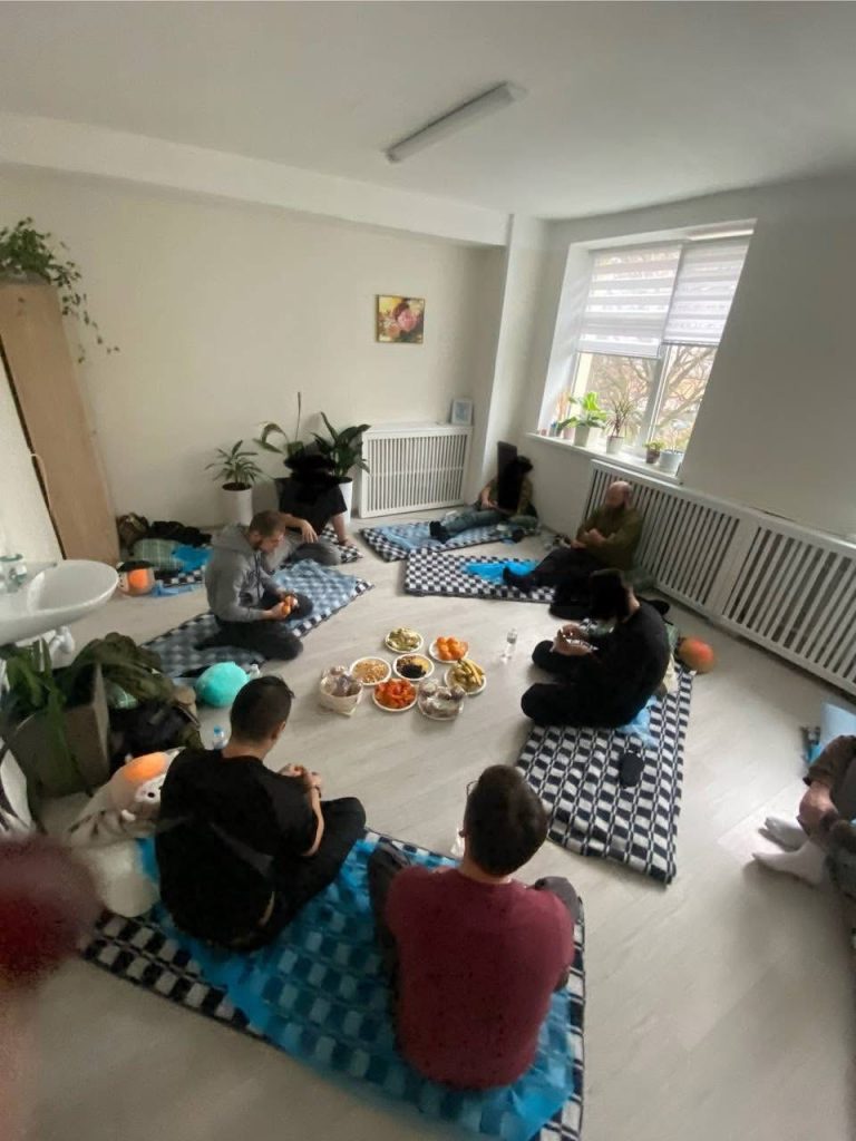 Treatment room. The veterans are sharing a meal together on the floor, sitting on their mattresses. Some food (fruit, vegetables...) is placed on plates in the middle of the room.